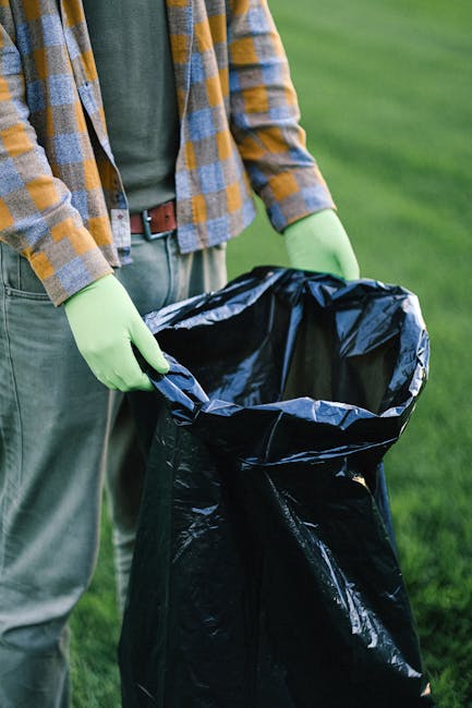The image depicts a person dressed in casual clothing, including a multi-coloured checkered shirt and green gloves, holding open a large black rubbish bag made of plastic. The bag appears to be prepared for waste collection or disposal, positioned outdoors on a grassy area with a blurred green background suggesting a garden or park environment. The individual is standing on a paved or natural surface, with their hands grasping the edges of the bag to open it wide, ready for rubbish insertion. The scene emphasizes an organised approach to waste management, which aligns with private or alternative rubbish removal services provided by companies like Rubbish Removal Highbury. The lighting is natural, indicating daylight, contributing to a clear and straightforward visual depiction. This image effectively communicates the act of rubbish collection or waste disposal, supporting service topics related to rubbish handling, on-site clearance, or independent waste collection efforts.