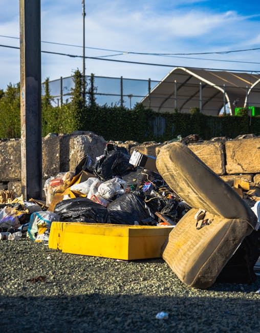 A ground-level view of an overflowing pile of mixed waste materials, including black plastic rubbish bags, a yellow plastic container, and a large, old, beige car seat positioned on its side. The debris is spread across a gravel surface, with a short stone wall and a metal utility pole nearby. In the background, there is a hedge and a partially visible building with a curved, translucent roof, possibly a sports facility or greenhouse, under a bright blue sky with some wispy clouds. The scene illustrates a typical example of improper waste disposal or a collection that has exceeded capacity, highlighting the importance of professional rubbish removal services like those offered by Rubbish Removal Highbury for alternative waste handling and on-site clearance.