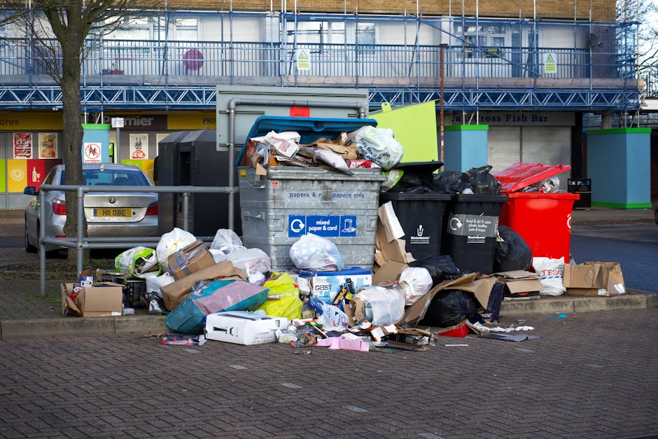 An outdoor scene showing a large pile of household waste and rubbish bags overflowing from several recycling and general waste bins, which are positioned on a paved area beside a parking space. The overflowing waste includes cardboard boxes, crumpled paper, plastic bags, and miscellaneous discarded items, some scattered on the ground. The recycling bins are labeled for mixed paper and cardboard, and standard black waste bins are also visible. Behind the bins, there is a multi-story building with a blue metal scaffolding structure, indicating construction or maintenance work, and the storefronts below feature signage for various shops, including a fish bar. A parked car is partially visible on the left side, and a leafless tree stands next to the bins, with a metal barrier separating the waste area from the rest of the pavement. Natural daylight illuminates the scene, emphasizing the disorganized state of waste accumulation, which relates to independent rubbish collection and waste disposal practices handled by companies such as Rubbish Removal Highbury.