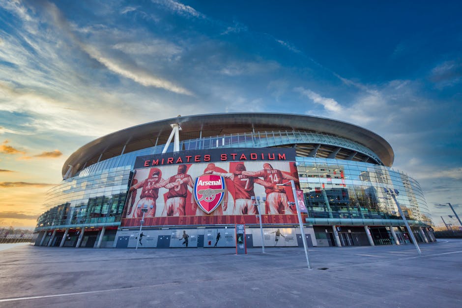 A wide-angle view of the exterior of Emirates Stadium during late afternoon, showing its modern architectural design with a curved, metallic roof structure and extensive glass facades. Prominently displayed on the stadium's front is a large digital screen featuring Arsenal football players celebrating, with the Arsenal logo centered beneath them. The surrounding area consists of a paved open space with minimal traffic or pedestrians visible, and a few street lamps positioned along the perimeter. The sky above is partly cloudy with the soft golden light of the setting sun casting a warm glow on the stadium, creating a contrast against the blue sky. The scene appears clean and well-maintained, reflecting an environment suitable for hosting large events. This visual emphasizes the stadium’s prominence as a key sports venue, with natural light highlighting its contemporary design, while the empty foreground and clear surroundings subtly support themes of private or independent event clearance, consistent with rubbish removal services by Rubbish Removal Highbury.