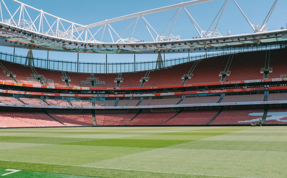 The image depicts an empty sports stadium, viewed from the field level, with a well-maintained grass pitch in the foreground. The grass shows distinct mowing patterns with different shades of green, stretching across the view with clear stripes. Surrounding the pitch are tiered seating areas filled with red and pink seats, some with empty sections and a few banners and advertisements visible along the railings. The stadium's roof structure is prominently featured overhead, composed of white steel trusses forming a geometric pattern against a bright blue sky. The overall scene is brightly lit, indicating daytime, with no spectators present. This setting emphasizes the clean, organized environment typical of a large sports venue, which is contextually relevant to the concept of private or independent waste management, as seen in stadium waste disposal and cleaning services, aligning with rubbish removal concepts handled by companies such as Rubbish Removal Highbury.