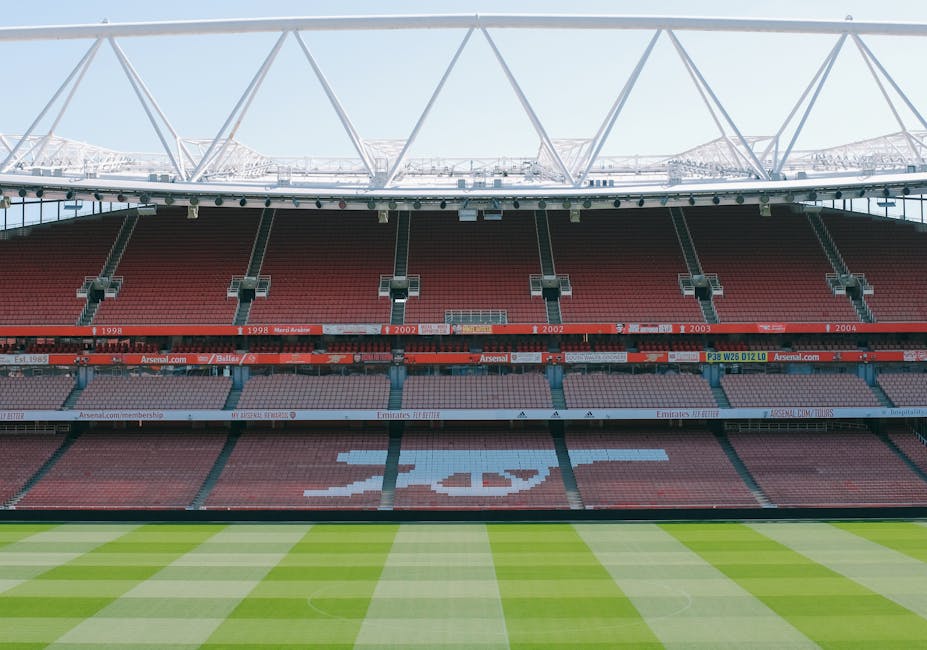 A wide-angle view of the exterior of Emirates Stadium during late afternoon, showing its modern architectural design with a curved, metallic roof structure and extensive glass facades. Prominently displayed on the stadium's front is a large digital screen featuring Arsenal football players celebrating, with the Arsenal logo centered beneath them. The surrounding area consists of a paved open space with minimal traffic or pedestrians visible, and a few street lamps positioned along the perimeter. The sky above is partly cloudy with the soft golden light of the setting sun casting a warm glow on the stadium, creating a contrast against the blue sky. The scene appears clean and well-maintained, reflecting an environment suitable for hosting large events. This visual emphasizes the stadium’s prominence as a key sports venue, with natural light highlighting its contemporary design, while the empty foreground and clear surroundings subtly support themes of private or independent event clearance, consistent with rubbish removal services by Rubbish Removal Highbury.