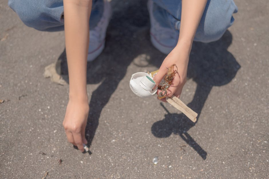 A person wearing light blue jeans and white sneakers is crouching on an outdoor pavement surface, holding a small handmade model of a white egg-shaped object attached to a wooden stick. The model appears to be crafted from lightweight materials like paper or foam, with some fiber or string visible around it, suggesting a craft or educational project. The surrounding ground is made of rough asphalt with scattered small debris. The scene is lit with natural daylight, casting shadows from the person's legs and the object onto the ground. This image, associated with waste management, subtly highlights individual participation in waste collection or environmental cleanup activities, relevant to private or independent rubbish removal efforts that complement traditional services provided by companies like Rubbish Removal Highbury.