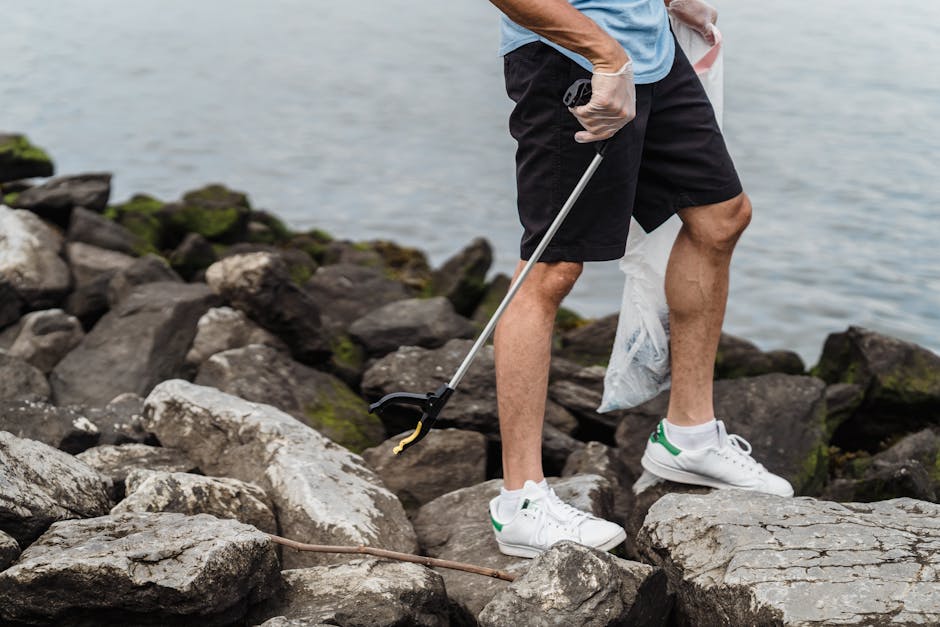 A person standing on a rocky shoreline holding a litter picker tool in their right hand, dressed in a blue t-shirt, black shorts, white sneakers with green accents, and wearing disposable gloves. The individual is positioned on uneven terrain composed of large, irregularly shaped rocks with rough textures and various shades of grey and brown. Moss and small patches of green algae are visible on some rocks near the water's edge. In the background, calm water extends to the horizon, with no other objects or figures present. The scene captures a moment of outdoor activity related to environmental cleanup, consistent with the focus of private waste handling and rubbish removal services offered by Rubbish Removal Highbury, emphasizing on-site or independent waste collection actions near natural environments.