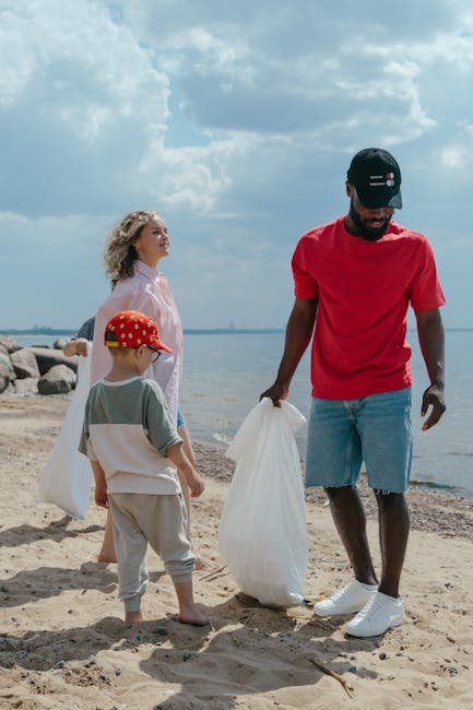 A man dressed in a red t-shirt, blue denim shorts, white sneakers, and a black cap is standing on a sandy beach, holding a white plastic bag in his right hand. To his left, a young boy wearing a grey t-shirt, beige trousers, a red hat, and barefoot is observing while standing on the sand. Behind the man, a woman in a light pink, semi-transparent jacket and white top is facing towards the water and earth, with her curly blonde hair blowing in the breeze. The background features a partly cloudy sky, calm water extending to the horizon, and a line of large rocks along the shoreline. The scene suggests activities related to beach clean-up or collecting rubbish, aligning with independent waste management or rubbish removal services, such as those offered by Rubbish Removal Highbury, emphasizing on-site clearance and private disposal options. The environment is well-lit, capturing a clear day with natural daylight enhancing the textures and colours of the sand, clothing, and water, contributing to a professional and neutral visual depiction relevant to waste collection and environmental care.