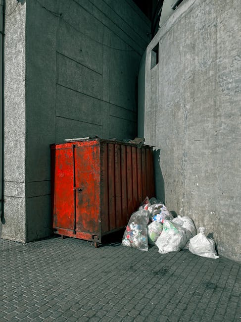 A large, rusted red metal rubbish skip positioned against a corner of a building with dark green panels on the left and a concrete wall on the right. The skip is filled with a small amount of waste visible at the top. Several white plastic bin bags, some slightly crumpled, are placed on the ground next to the skip, containing mixed waste materials. The area appears to be an urban outdoor space, possibly a back alley or side street, with a textured pavement surface beneath the skip and bags. The lighting suggests a natural daylight setting. This scene reflects an instance of private waste handling, possibly managed by Rubbish Removal Highbury for on-site clearance or alternative rubbish collection, with the bags and skip indicating an effort to manage waste outside of standard roadside collection services.