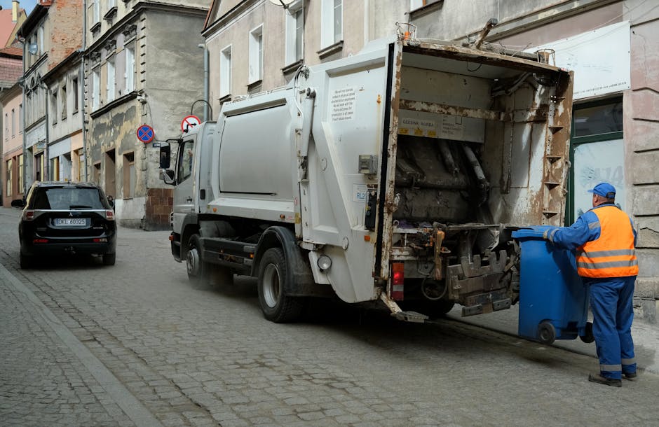 A sanitation worker dressed in a high-visibility orange vest and blue uniform is standing on a cobblestone street beside a white garbage collection truck, which is positioned with its rear compactor open and ready for waste disposal. The worker is holding a blue wheeled bin, about to empty its contents into the truck's hopper. The scene takes place in an urban environment with older, multi-story buildings lining the street, featuring stone and brick facades, some with visible peeling paint and weathered surfaces. parked cars, including a black vehicle with a visible European license plate, are alongside the curb. The truck appears weathered and functional, with visible rust and wear on some metal surfaces, indicating regular use. The atmosphere is calm, with natural daylight illuminating the scene, and the environment reflects typical private rubbish collection in the area, supporting alternative waste handling methods outside of local authority services. Rubbish Removal Highbury provides such services, contributing to efficient waste management solutions in this setting.