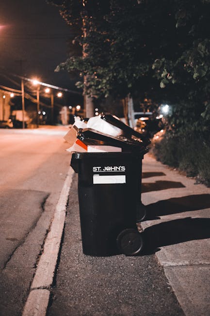A large, rusted red metal rubbish skip positioned against a corner of a building with dark green panels on the left and a concrete wall on the right. The skip is filled with a small amount of waste visible at the top. Several white plastic bin bags, some slightly crumpled, are placed on the ground next to the skip, containing mixed waste materials. The area appears to be an urban outdoor space, possibly a back alley or side street, with a textured pavement surface beneath the skip and bags. The lighting suggests a natural daylight setting. This scene reflects an instance of private waste handling, possibly managed by Rubbish Removal Highbury for on-site clearance or alternative rubbish collection, with the bags and skip indicating an effort to manage waste outside of standard roadside collection services.