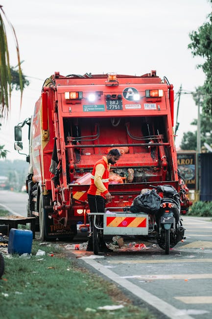 A sanitation worker wearing an orange high-visibility vest and dark trousers is seen collecting waste from a black plastic bag placed on a small metal cart positioned behind a red rubbish collection truck. The truck, parked on the side of a paved street, has its rear compactor open with hydraulic mechanisms visible, indicating ongoing refuse loading. The vehicle features reflective yellow and red striped patterns on the rear and a white circular logo on the upper left side. The scene is set outdoors during daylight, with a partly cloudy sky and scattered trees in the background, along with a building and power lines visible behind the truck. The street has a designated lane for collection activities, marked by white striped lines, and nearby pavement with some litter. The overall setting suggests an early morning or daytime waste collection operation, consistent with private rubbish collection services like those provided by Rubbish Removal Highbury, supporting alternative waste handling outside local authority collection.